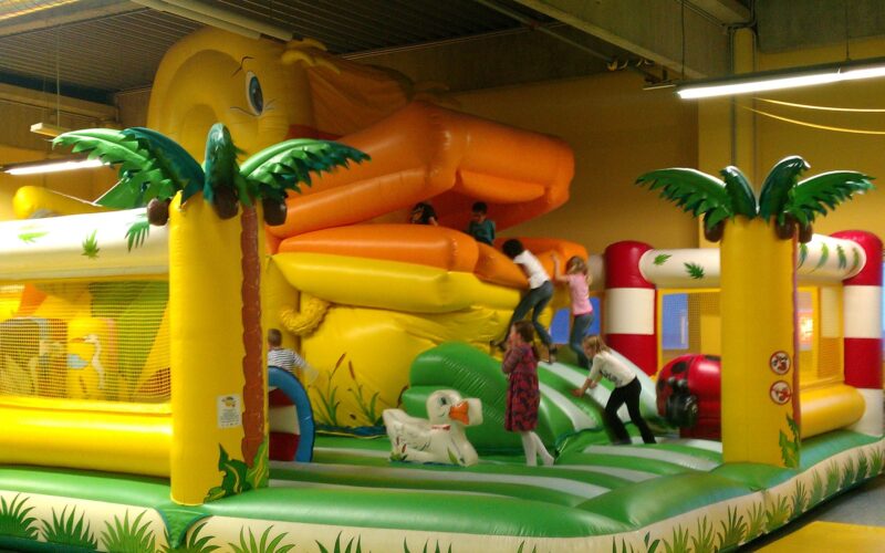 Kinder spielen in einer aufblasbaren Dschungel-Hüpfburg mit Palmen und einer großen gelben Rutsche.