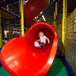 Ein Kind rutscht eine große rote Röhrenrutsche in einem Indoor-Spielplatz mit Sicherheitsnetzen und buntem Bodenbelag hinunter.
