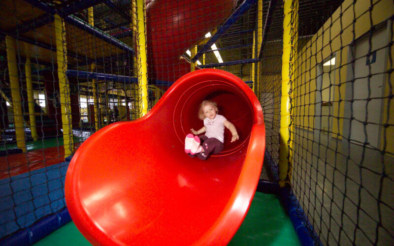 Ein Kind rutscht eine große rote Röhrenrutsche in einem Indoor-Spielplatz mit Sicherheitsnetzen und buntem Bodenbelag hinunter.