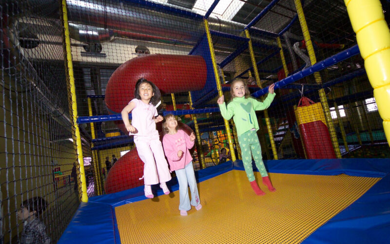 Drei Kinder springen auf einem gelben Trampolin in einem Indoor-Spielplatz mit Netzwänden und einer roten Rutsche im Hintergrund.