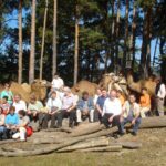 Eine Gruppe von Menschen sitzt auf Baumstämmen in einem bewaldeten Gebiet mit mehreren Kamelen und Bäumen im Hintergrund.