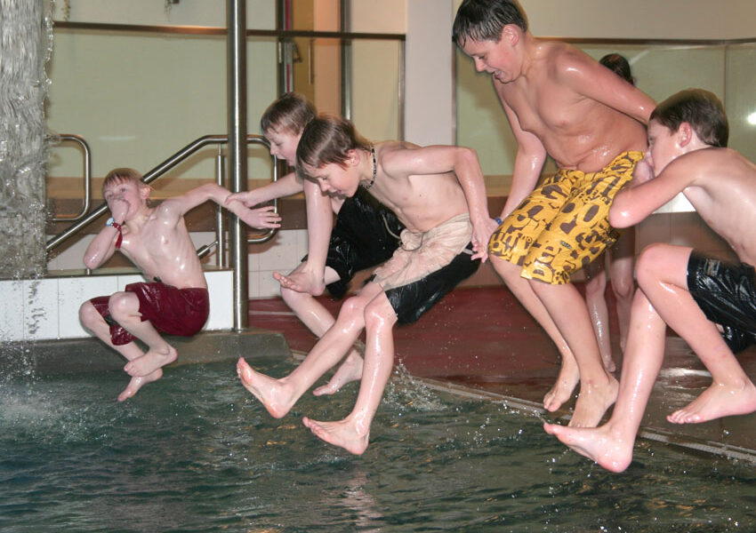 Vier Jungen in Badehosen springen gemeinsam in ein Hallenbad, mitten in der Luft, und das Wasser spritzt um sie herum.