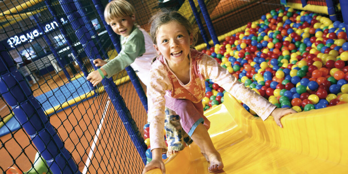 Zwei Kinder spielen auf einer gelben Rutsche neben einer farbenfrohen Ballgrube in einem Indoor-Spielbereich und lächeln, während sie klettern.