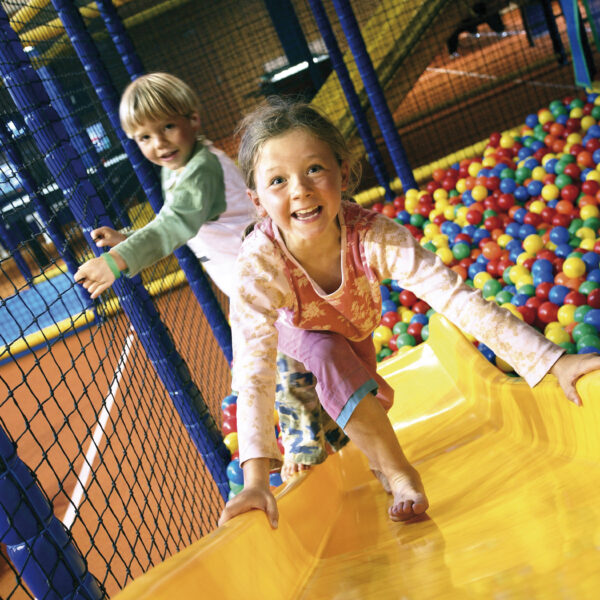 Zwei Kinder spielen auf einer gelben Rutsche neben einer farbenfrohen Ballgrube in einem Indoor-Spielbereich und lächeln, während sie klettern.