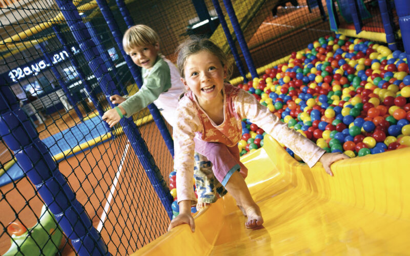 Zwei Kinder spielen auf einer gelben Rutsche neben einer farbenfrohen Ballgrube in einem Indoor-Spielbereich und lächeln, während sie klettern.
