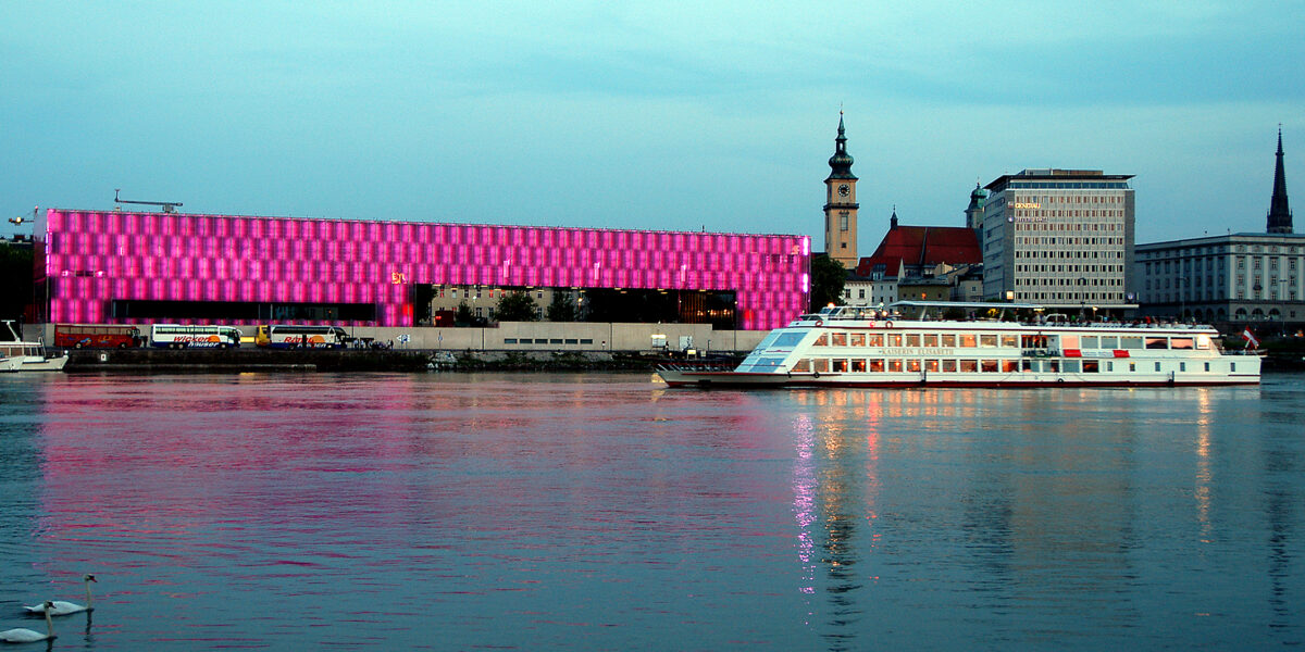 Ein modernes Flussschiff fährt an einem hell erleuchteten rosa Gebäude und einer Stadtlandschaft vorbei, die sich in der Abenddämmerung im ruhigen Wasser spiegelt.
