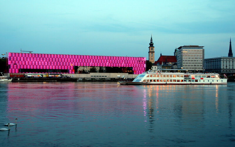 Ein modernes Flussschiff fährt an einem hell erleuchteten rosa Gebäude und einer Stadtlandschaft vorbei, die sich in der Abenddämmerung im ruhigen Wasser spiegelt.