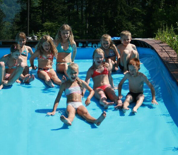 Eine Gruppe von Kindern in Badeanzügen sitzt an einem sonnigen Tag im Freien, umgeben von Bäumen und Grün, am Rande eines Pools mit Wasser.
