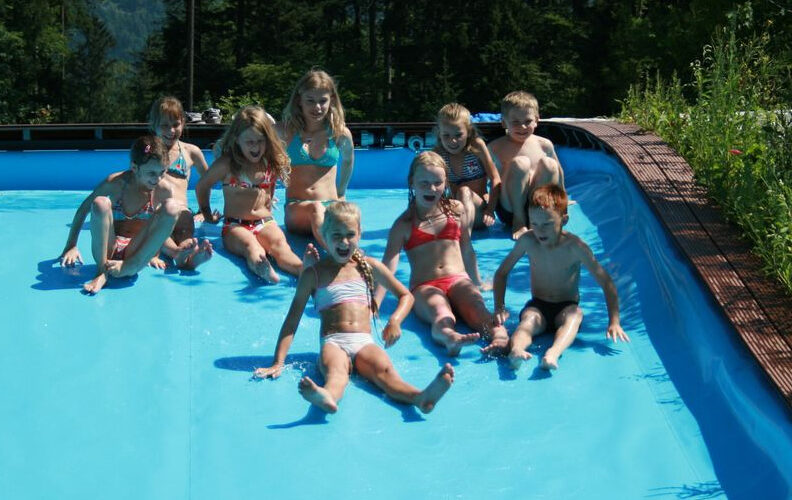 Eine Gruppe von Kindern in Badeanzügen sitzt an einem sonnigen Tag im Freien, umgeben von Bäumen und Grün, am Rande eines Pools mit Wasser.