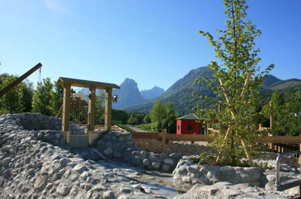 Ein aus Stein gebauter Wasserspielplatz mit Holzstrukturen, einem kleinen roten Gebäude, Bäumen und Bergen im Hintergrund unter einem klaren Himmel.