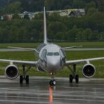 Ein Verkehrsflugzeug mit einem aufgemalten Tiergesicht auf der Nase rollt auf einer nassen Landebahn, mit Bäumen und Gebäuden im Hintergrund.