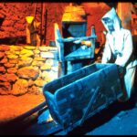 Eine als Bergmann verkleidete Schaufensterpuppe steht neben einem Holzkarren in einer schwach beleuchteten Bergbauausstellung mit Steinwänden.