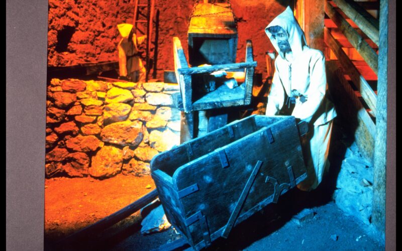 Eine als Bergmann verkleidete Schaufensterpuppe steht neben einem Holzkarren in einer schwach beleuchteten Bergbauausstellung mit Steinwänden.