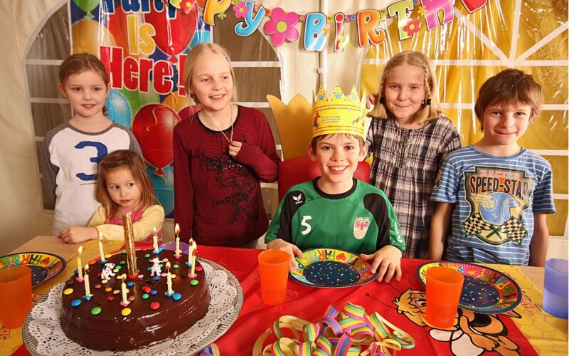 Sechs Kinder lächeln um einen Geburtstagstisch mit einem Schokoladenkuchen, Kerzen, bunten Tellern, Tassen und einem "Happy Birthday"-Banner. Ein Kind trägt eine Papierkrone.