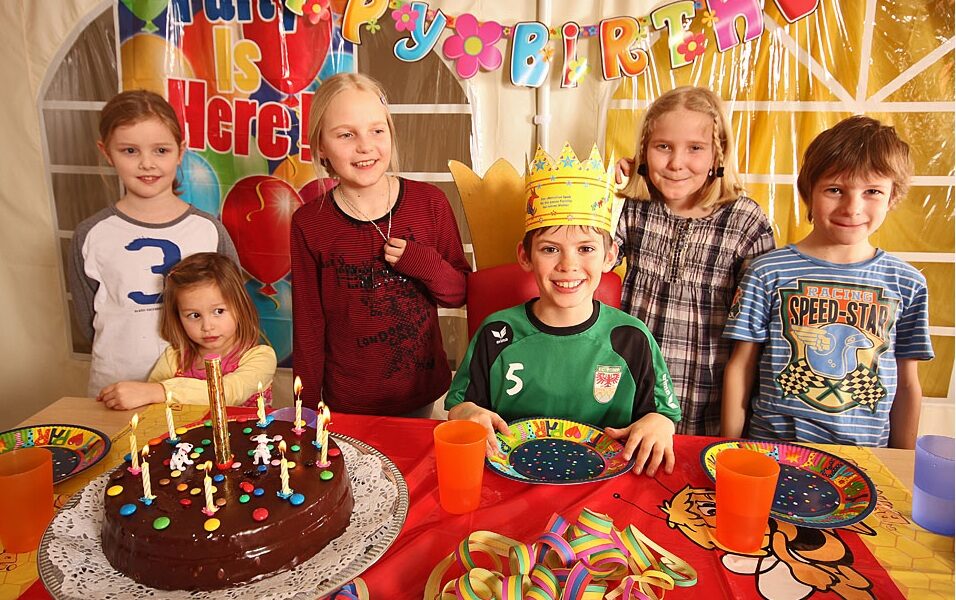 Sechs Kinder lächeln um einen Geburtstagstisch mit einem Schokoladenkuchen, Kerzen, bunten Tellern, Tassen und einem "Happy Birthday"-Banner. Ein Kind trägt eine Papierkrone.