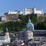 Ein Blick auf die Festung Hohensalzburg auf einer Anhöhe über dem barocken Dom und den Gebäuden der Stadt Salzburg unter blauem Himmel.