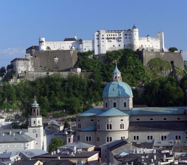 Ein Blick auf die Festung Hohensalzburg auf einer Anhöhe über dem barocken Dom und den Gebäuden der Stadt Salzburg unter blauem Himmel.