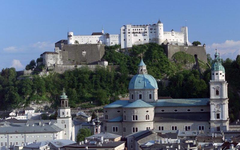 Ein Blick auf die Festung Hohensalzburg auf einer Anhöhe über dem barocken Dom und den Gebäuden der Stadt Salzburg unter blauem Himmel.