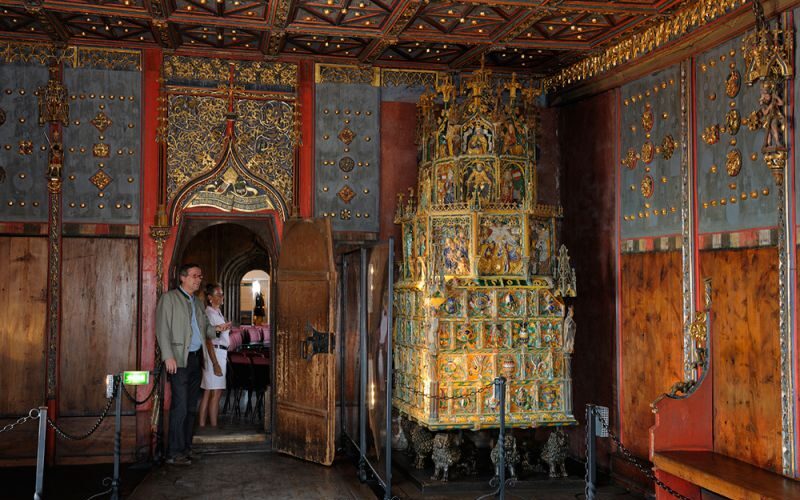 Zwei Personen stehen in einem reich verzierten Raum mit kunstvollen Goldverzierungen, Holzwänden und einem großen, dekorativen Keramikofen.