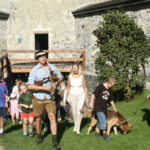 Eine Gruppe von Erwachsenen und Kindern, einige mit Ziegen an der Leine, spaziert an einem sonnigen Tag auf einem grasbewachsenen Weg in der Nähe eines Steinhauses.