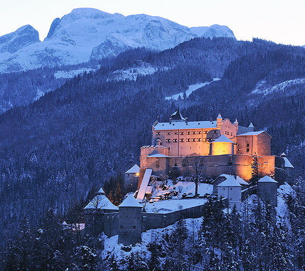 Ein großes, in der Abenddämmerung beleuchtetes Schloss steht auf einem verschneiten, bewaldeten Hügel, mit hohen, schneebedeckten Bergen im Hintergrund.