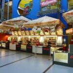 Ein hell erleuchteter Kiosk in einem Kino bietet Popcorn, Snacks und Getränke an. Darüber sind Menütafeln und das Coca-Cola-Logo zu sehen.
