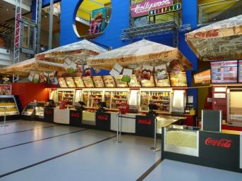 Ein hell erleuchteter Kiosk in einem Kino bietet Popcorn, Snacks und Getränke an. Darüber sind Menütafeln und das Coca-Cola-Logo zu sehen.