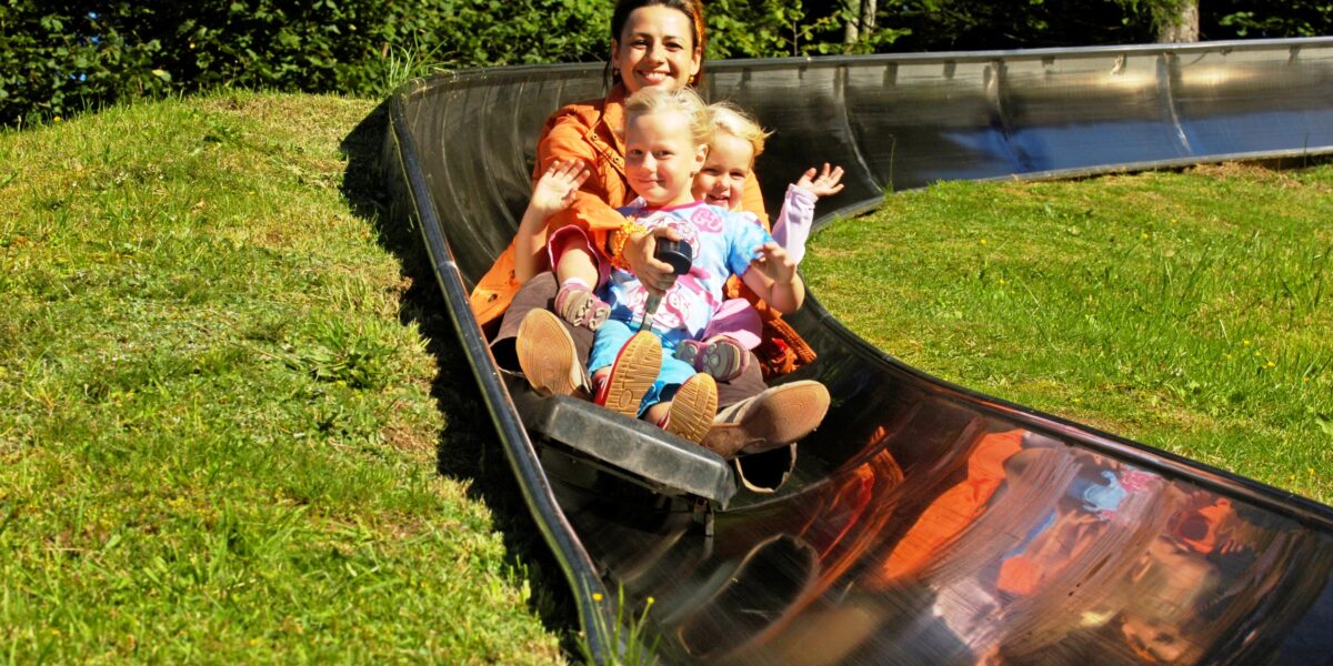Ein Erwachsener und zwei Kinder fahren gemeinsam auf einer Metallrutsche im Freien, umgeben von grünem Gras und Bäumen.