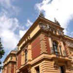 Reich verziertes historisches Gebäude mit Akzenten aus rotem Backstein und Stein, dekorativen Skulpturen, Bogenfenstern und einem bewölkten blauen Himmel im Hintergrund.