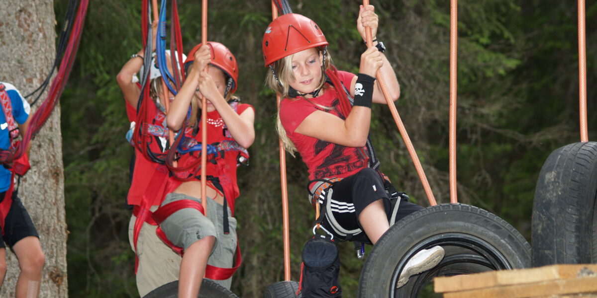 Kinder, die Helme und Klettergurte tragen, navigieren durch einen Hochseilgarten im Freien und treten auf hängende Reifen, die an Seilen in einem Waldgebiet aufgehängt sind.