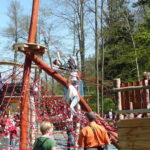 Kinder klettern und spielen auf einer großen Seilstruktur und einem Holzspielplatz im Freien, umgeben von Erwachsenen und Bäumen.