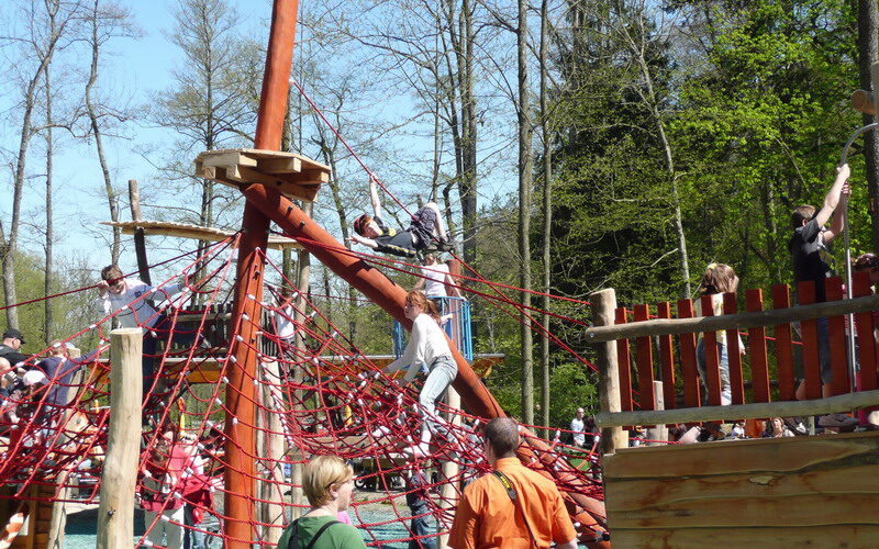 Kinder klettern und spielen auf einer großen Seilstruktur und einem Holzspielplatz im Freien, umgeben von Erwachsenen und Bäumen.