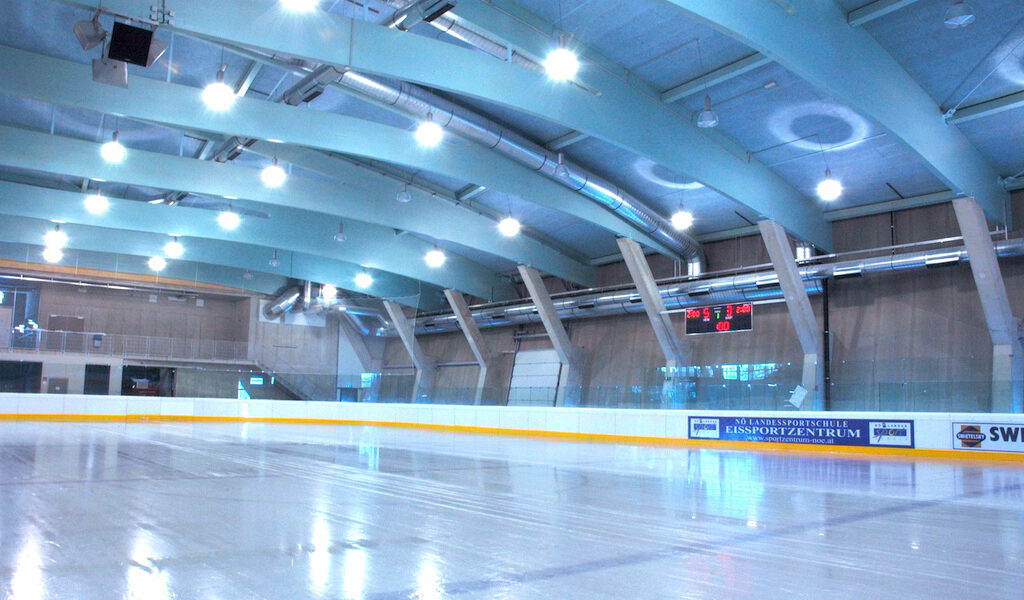 Eislaufhalle mit glattem, reflektierendem Eis, leeren Tribünen, Oberlicht und einer digitalen Anzeigetafel, die die Spielstände an der hinteren Wand anzeigt.
