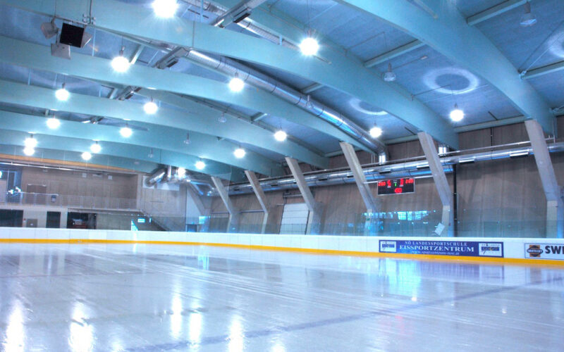 Eislaufhalle mit glattem, reflektierendem Eis, leeren Tribünen, Oberlicht und einer digitalen Anzeigetafel, die die Spielstände an der hinteren Wand anzeigt.