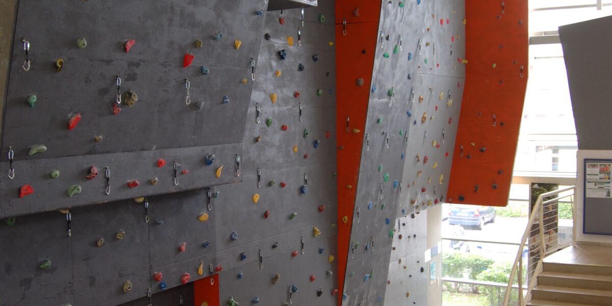 Indoor-Kletterwand mit verschiedenfarbigen Griffen und Sicherheitsmatten am Fuß, die sich neben einer Treppe in einer modernen Anlage befindet.