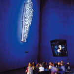 Eine Gruppe von Kindern sitzt auf dem Boden und betrachtet eine beleuchtete, abstrakte Kunstinstallation, die hoch oben an einer blauen Wand in einem schwach beleuchteten Raum angebracht ist.