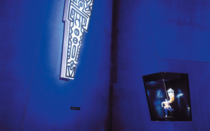 Eine Gruppe von Kindern sitzt auf dem Boden und betrachtet eine beleuchtete, abstrakte Kunstinstallation, die hoch oben an einer blauen Wand in einem schwach beleuchteten Raum angebracht ist.