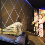 Zwei Mädchen betrachten eine große, verzierte Federballskulptur, die auf einem Kissen in einer Museumsausstellung ausgestellt ist.