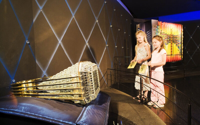 Zwei Mädchen betrachten eine große, verzierte Federballskulptur, die auf einem Kissen in einer Museumsausstellung ausgestellt ist.