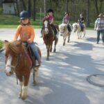 Vier Kinder reiten auf Ponys auf einem gepflasterten Weg, während ein Erwachsener in der Nähe spazieren geht und die Gruppe durch ein bewaldetes Parkgelände führt.