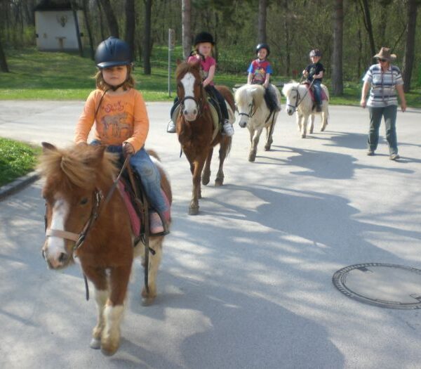 Vier Kinder reiten auf Ponys auf einem gepflasterten Weg, während ein Erwachsener in der Nähe spazieren geht und die Gruppe durch ein bewaldetes Parkgelände führt.