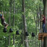 Zwei Personen nehmen an einem Hochseilgarten im Freien teil; eine überquert hängende schwarze Eimer, während die andere auf einer Holzplattform zwischen hohen Bäumen steht.