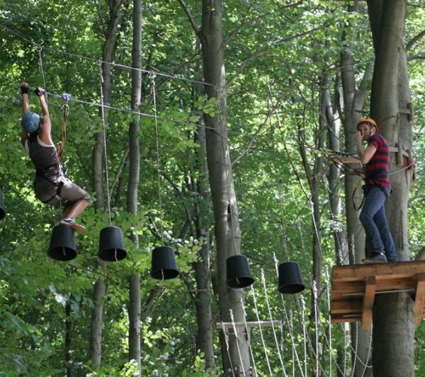 Zwei Personen nehmen an einem Hochseilgarten im Freien teil; eine überquert hängende schwarze Eimer, während die andere auf einer Holzplattform zwischen hohen Bäumen steht.
