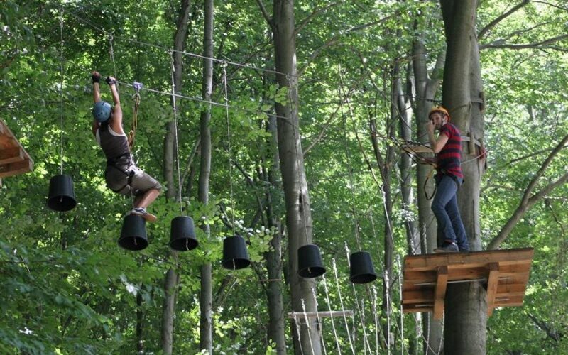 Zwei Personen nehmen an einem Hochseilgarten im Freien teil; eine überquert hängende schwarze Eimer, während die andere auf einer Holzplattform zwischen hohen Bäumen steht.
