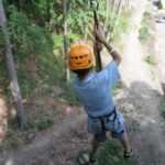 Eine Person, die einen Helm und ein Gurtzeug trägt, fährt auf einer Seilrutsche über einen unbefestigten Weg in einem Waldgebiet.