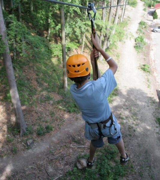 Eine Person, die einen Helm und ein Gurtzeug trägt, fährt auf einer Seilrutsche über einen unbefestigten Weg in einem Waldgebiet.