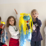 Vier kleine Kinder stehen drinnen und halten bunte, handgefertigte Kunstwerke mit Luftschlangen und Dekorationen in die Höhe und posieren vor einer schlichten weißen Wand.