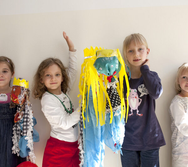 Vier kleine Kinder stehen drinnen und halten bunte, handgefertigte Kunstwerke mit Luftschlangen und Dekorationen in die Höhe und posieren vor einer schlichten weißen Wand.