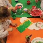 Die Kinder sitzen an einem Tisch und malen mit Pinseln auf Papier und Tellern, mit verschiedenen Farben, mit Farbbehältern und Malutensilien, die um sie herum verteilt sind.