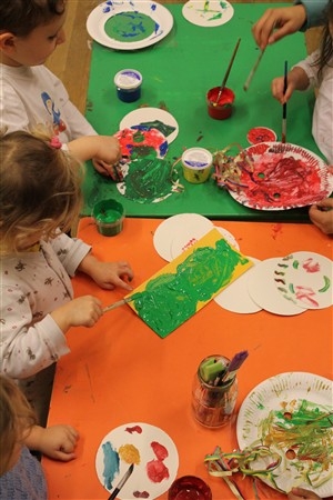 Die Kinder sitzen an einem Tisch und malen mit Pinseln auf Papier und Tellern, mit verschiedenen Farben, mit Farbbehältern und Malutensilien, die um sie herum verteilt sind.